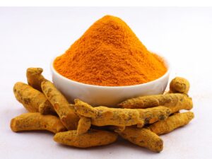 A mound of turmeric in a white bowl with some turmeric root in front of it.