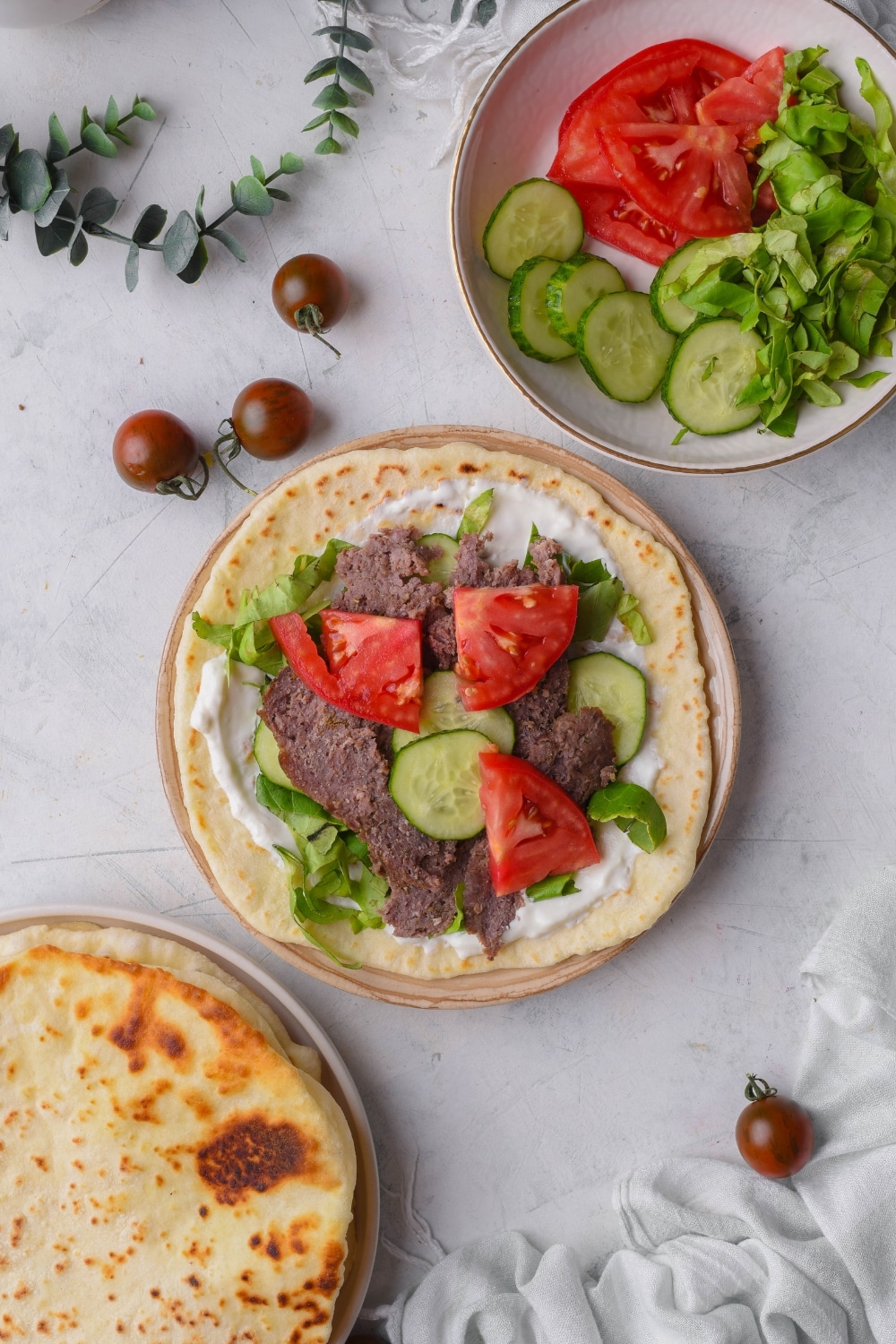 An open-face gyro with tzatziki sauce, lettuce, thinly sliced beef, cucumber, and tomato slices. The gyro is next to a bowl of veggies.