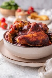 Part of a white bowl that is filled with bbq chicken wings. The bowl is on a white plate on a grey counter.
