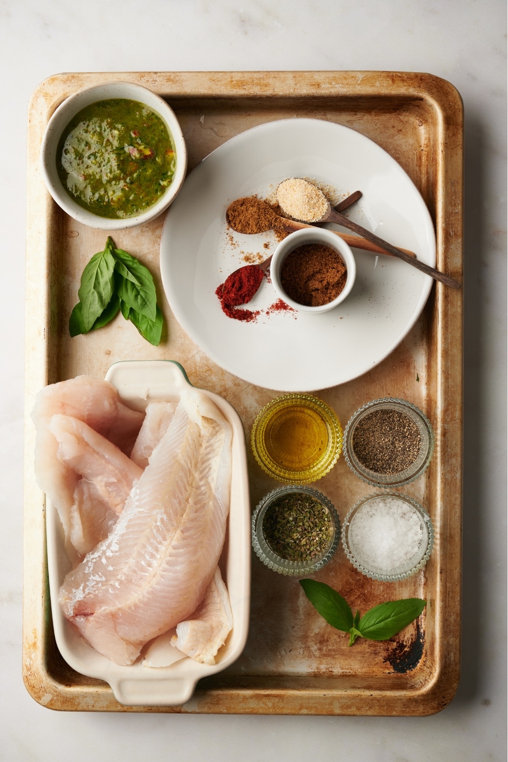 Catfish in a dish on a baking sheet, a small bowl of pepper, a small bowl of salt, a small bowl of oil, and a bunch of spices on a plate.