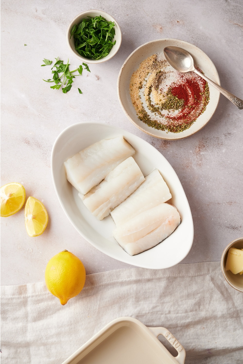 A bunch of seasonings in a bowl, four mahi mahi fillets on a white dish, and lemon wedges on a white counter.