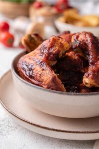 Part of a white bowl on a white plate with barbecue chicken wings in it.