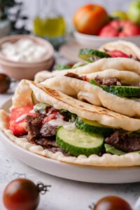 A gyros with thinly sliced meat, sliced cucumber, lettuce, tomato and tzatziki sauce.