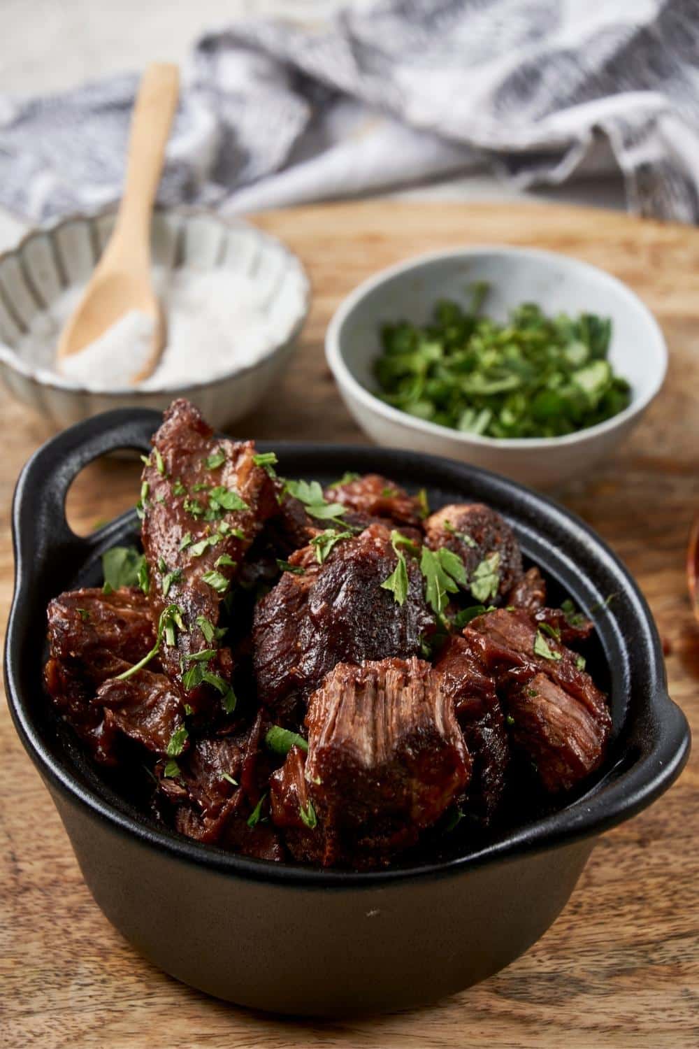 Burnt ends in a small cast iron bowl garnished with fresh parsley, next to small bowls of salt and parsley.