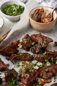 Burnt ends garnished with fresh parsley on a baking sheet, next to a small bowl of bbq sauce with a spoon and a bowl of chopped parsley.