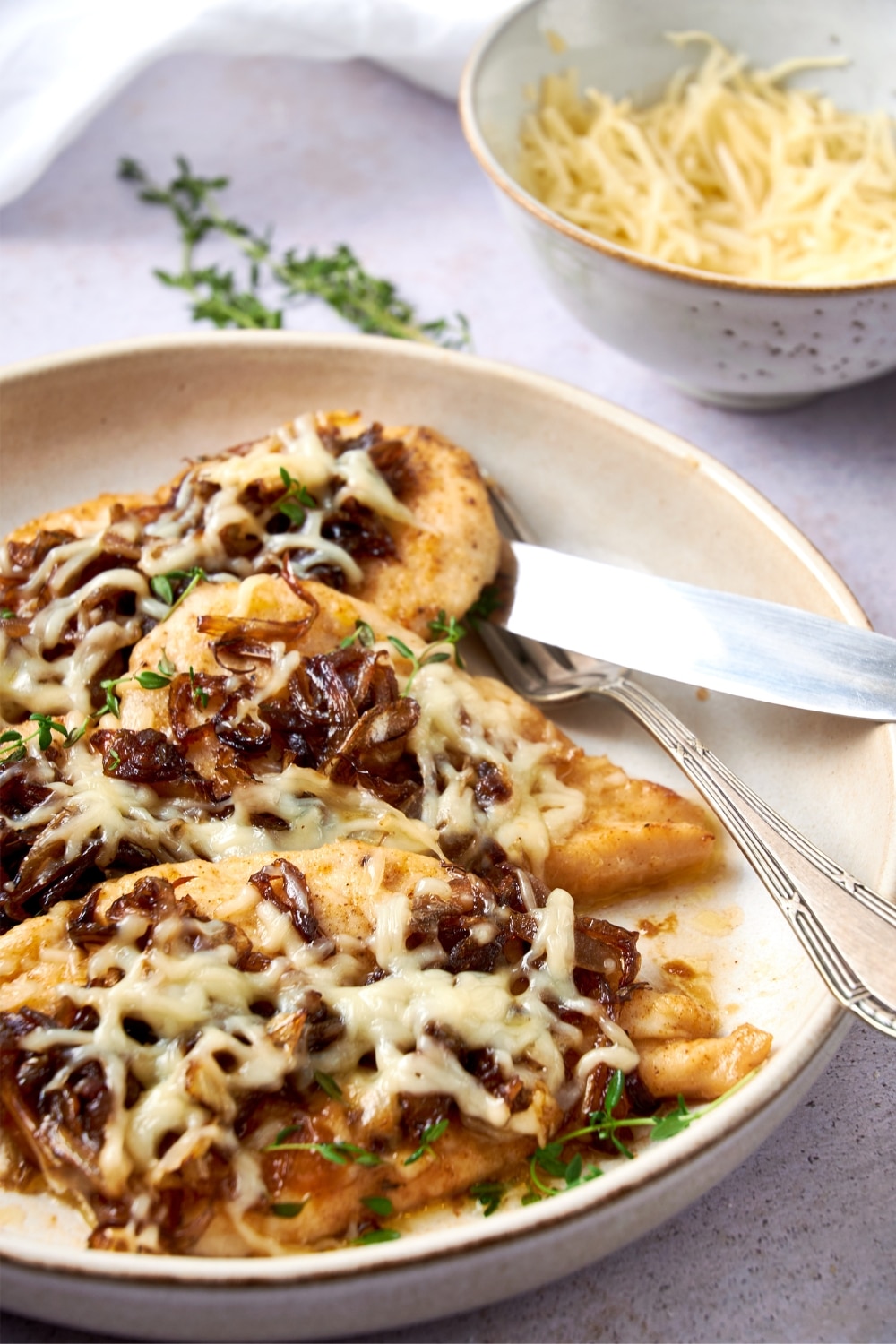 A plate of French onion chicken garnished with fresh thyme and served with a fork and knife.