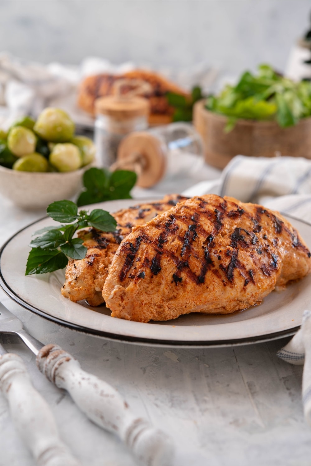 Grilled yogurt-marinated chicken breasts garnished with fresh herbs, next to bowls of salad greens and brussels sprouts and another plate of chicken.