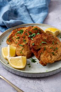 Three breaded turkey cutlets on a plate garnished with fresh herbs and served with lemon wedges.