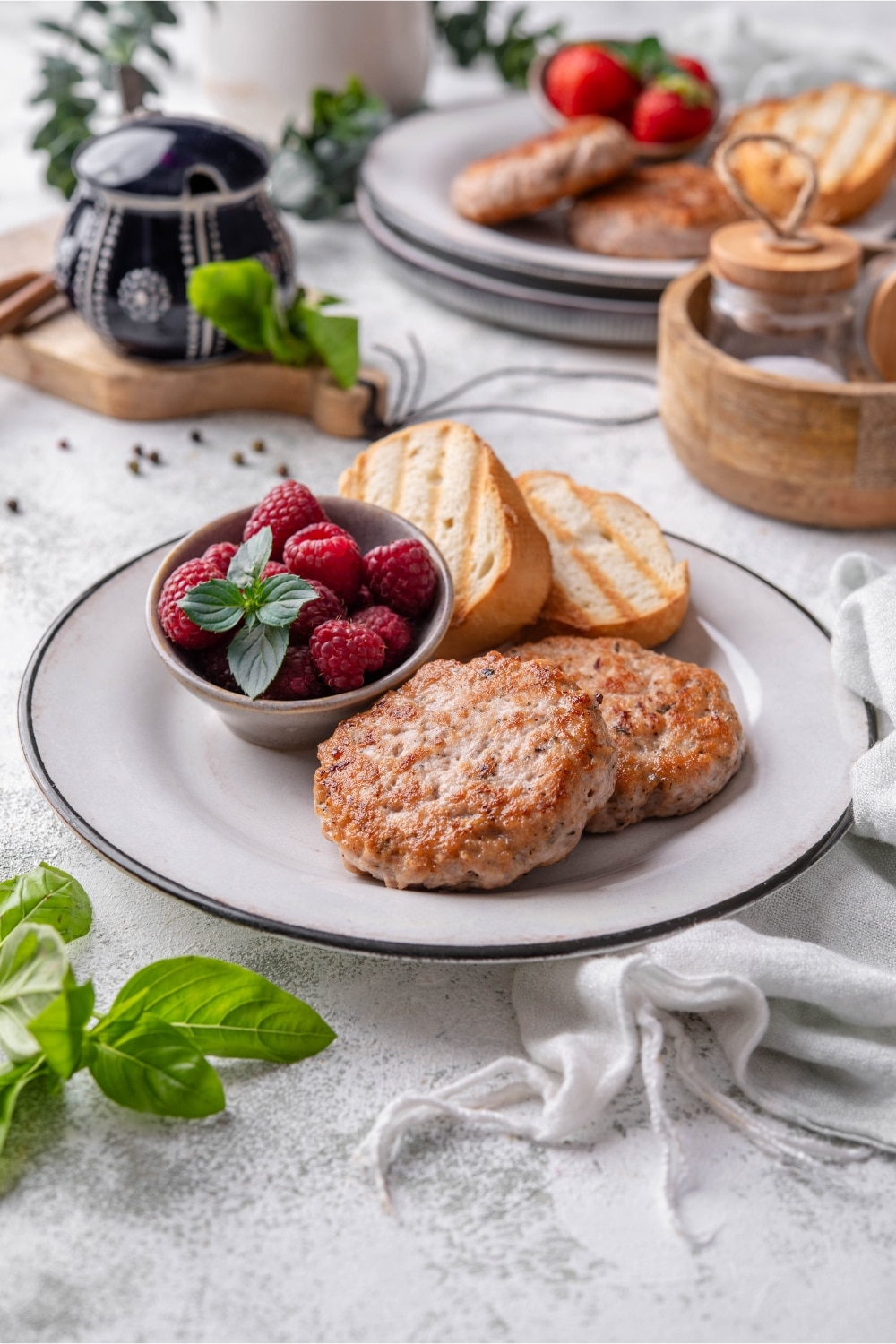 A turkey sausage breakfast plate with toasted baguette slices and raspberries. A second bowl served with strawberries can be seen in the back.