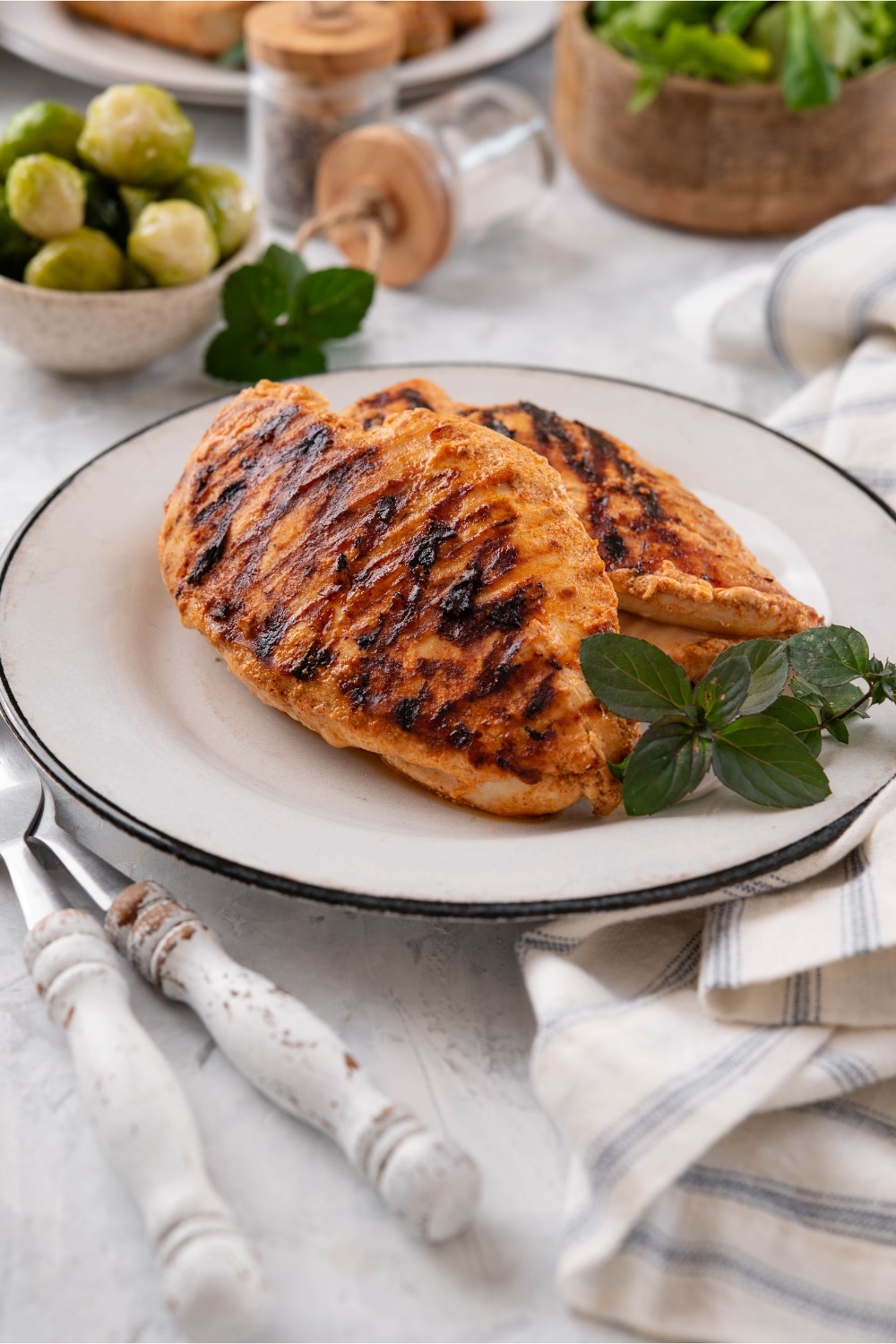 Grilled chicken breasts on a white plate, garnished with fresh herbs, next to bowls of salad greens and brussels sprouts.