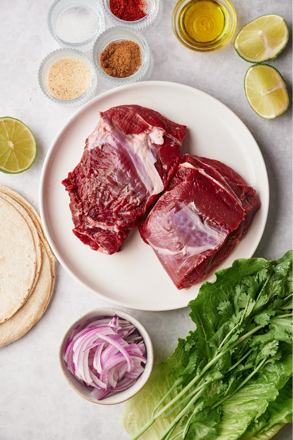 Two flank steaks on a plate, a bowl of onion powder, a bowl of re onions, a bowl of paprika, and a bowl of chili powder all on a white counter.