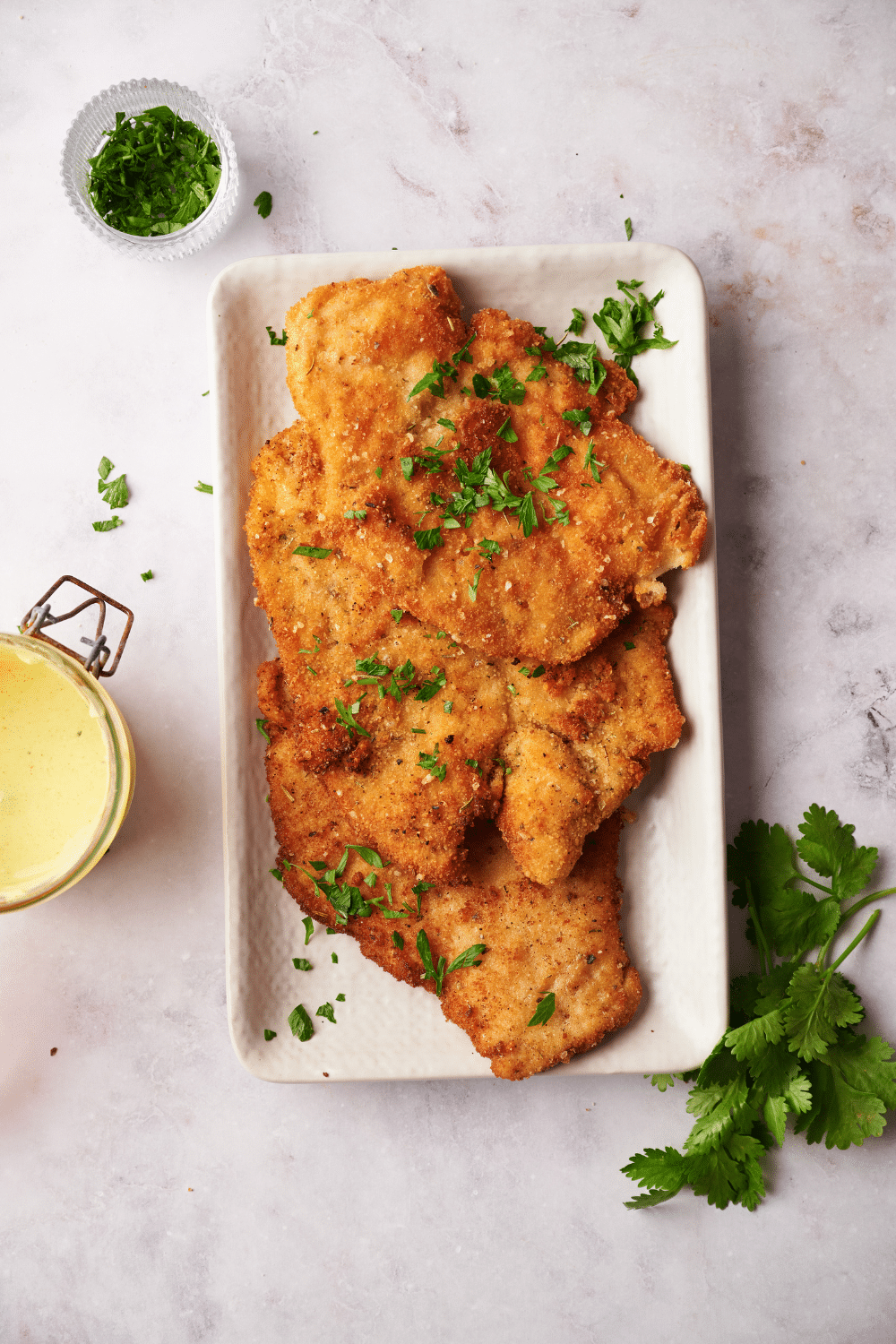 Herbs on top of three pieces of breaded chicken that are overlapping one another on top of a white rectangular baking dish on a white counter.