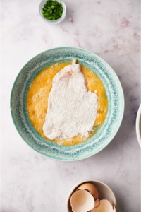 A blue bowl filled with a beaten egg mixture and a flour-coated chicken cutlet is in the bowl.