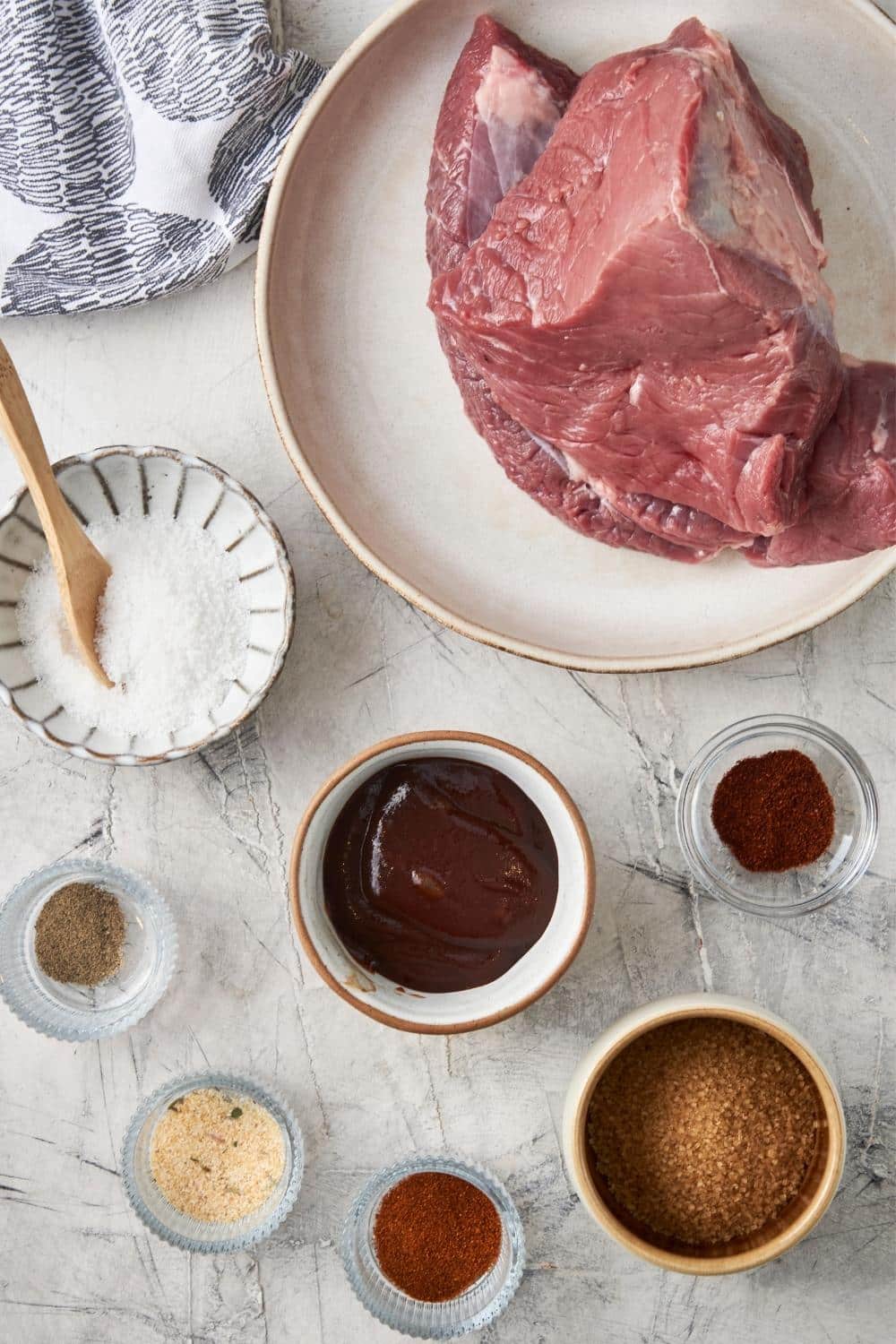 Chuck roast in a plate next to bowls of salt, spices, brown sugar, and bbq sauce.