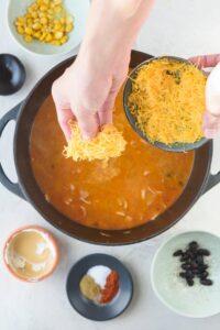Adding shredded cheddar cheese to a pot of chicken enchilada soup.