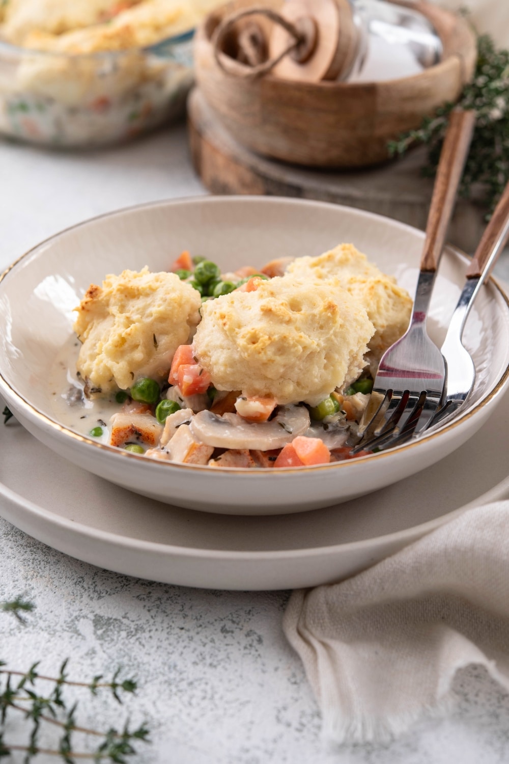 Three biscuits on top of chicken and veggies in a creamy sauce in a bowl with two forks. The bowl is on a white plate on a grey counter.