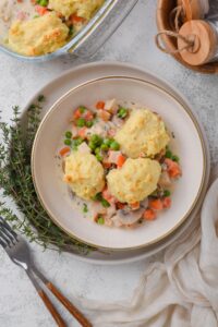 A top-down view of a white bowl full of chicken and biscuits casserole. Three biscuits sit atop the casserole filling. The bowl is sitting on a white plate with a sprig of fresh herbs.