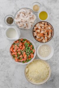 Sliced white mushrooms, chopped chicken, frozen carrots and peas, milk, flour, and biscuit mix are all in separate bowls on a white counter.