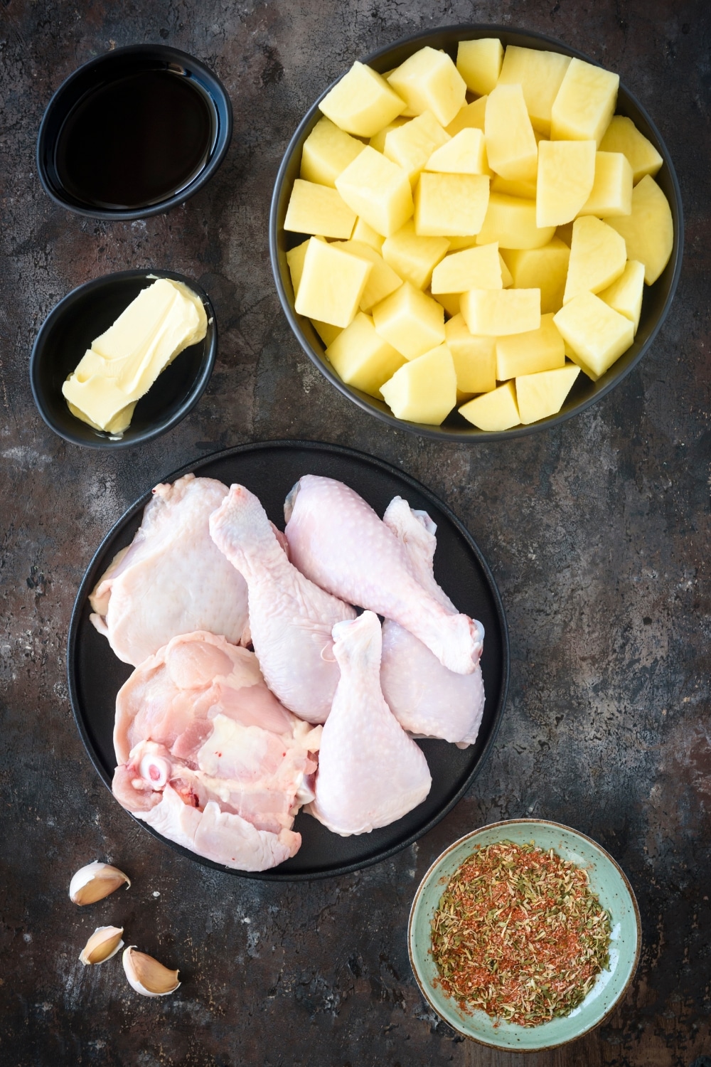 A bowl of potatoes, a plate filled with chicken, and a bowl of seasoning on a black counter.