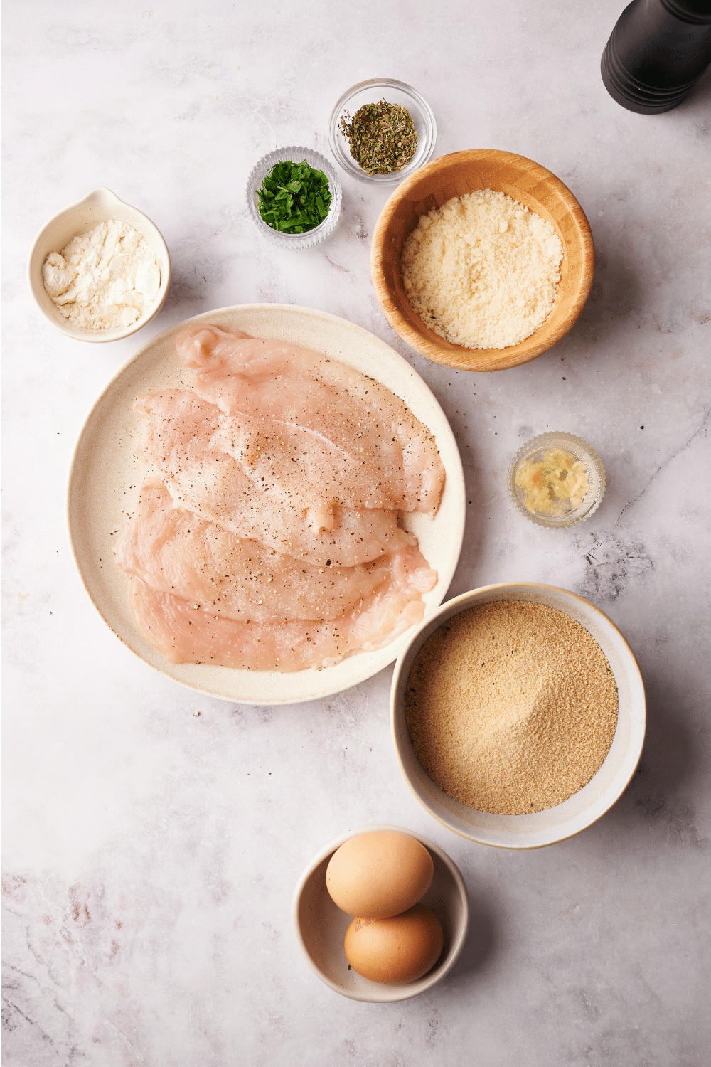 An assortment of ingredients including a plate of chicken cutlets and bowls of parmesan cheese, breadcrumbs, two eggs, flour, minced garlic, and seasonings.