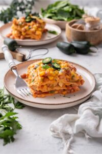 A square of enchilada casserole with laters of tortillas, corn, beans, cheese, and sauce.