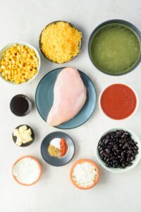 A plate of chicken breast and bowls of chicken broth, shredded cheddar cheese, corn kernels, olive oil, butter, cream cheese, spices, onions, black beans, and tomato sauce.