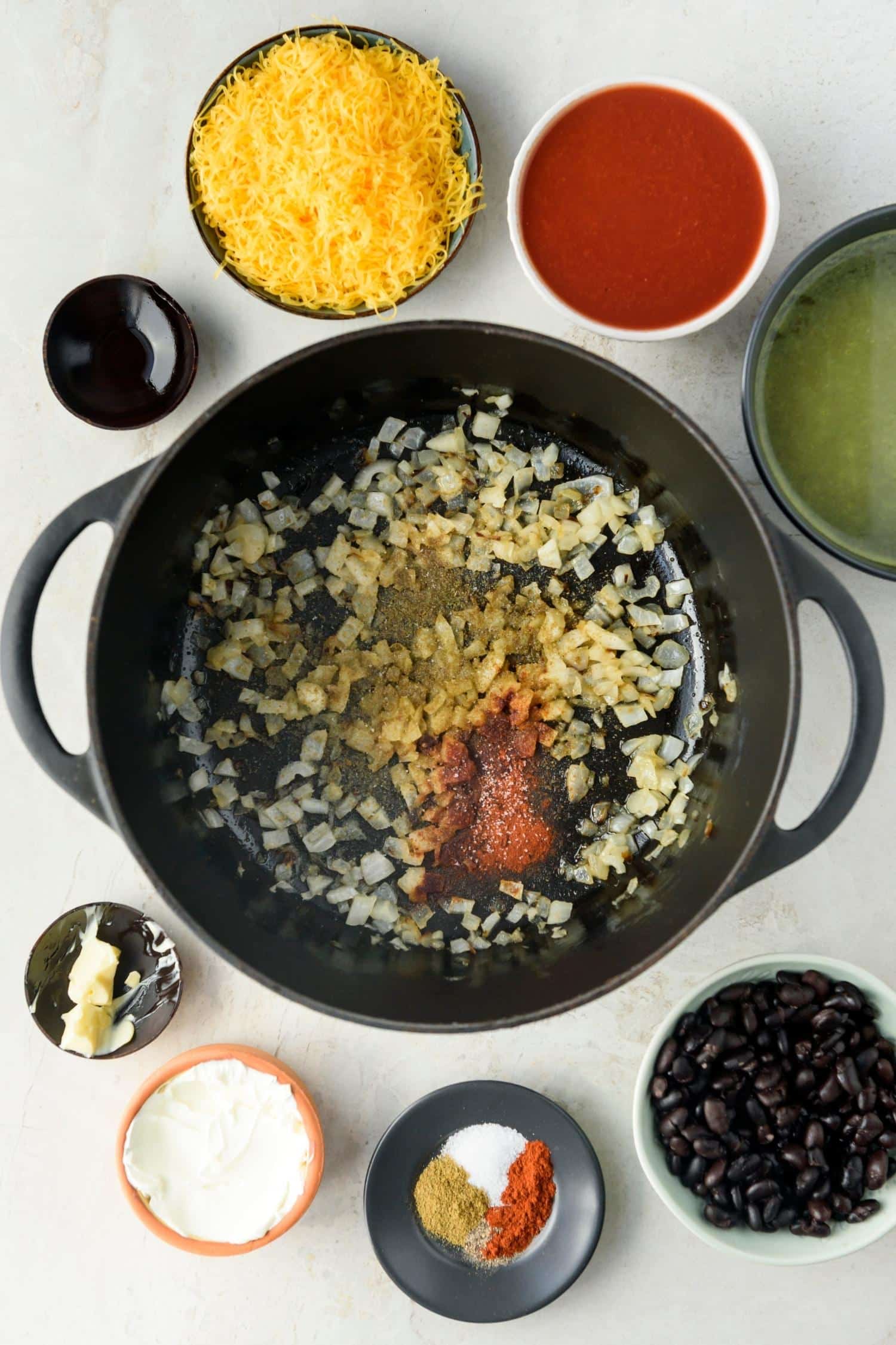 Chopped onions and spices in a large pot, surrounded by bowls of the rest of the ingredients.