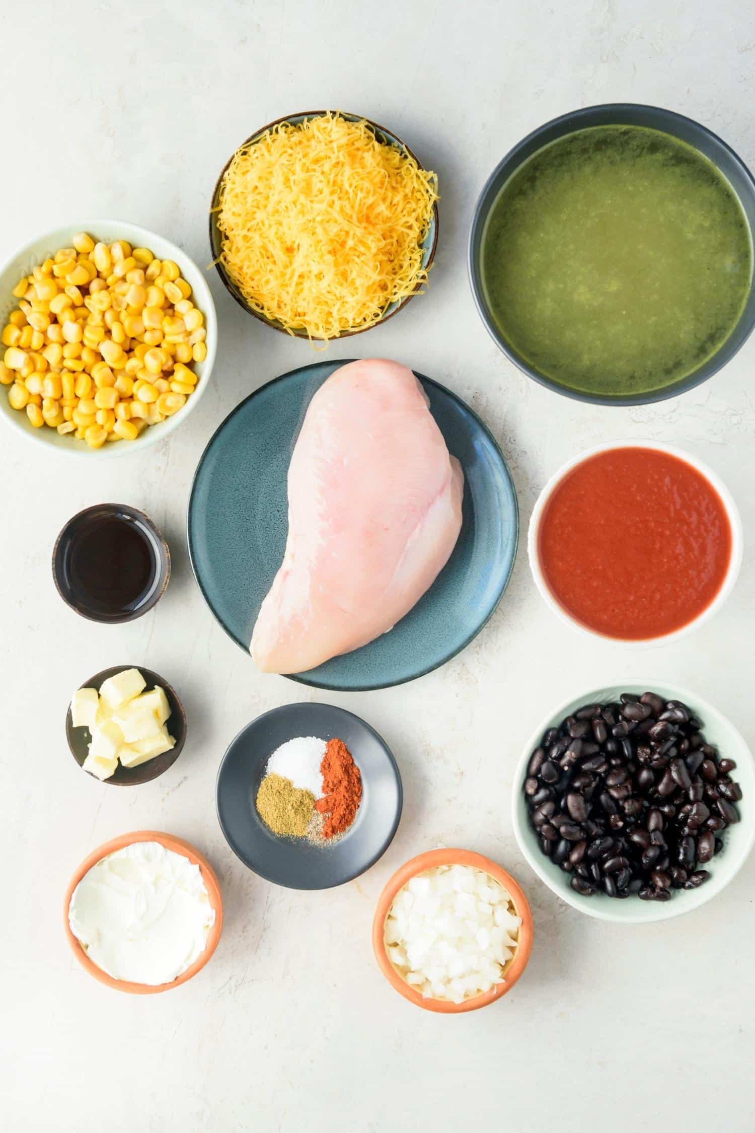 A plate of chicken breast and bowls of chicken broth, shredded cheddar cheese, corn kernels, olive oil, butter, cream cheese, spices, onions, black beans, and tomato sauce.