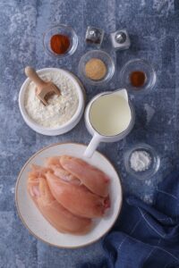 Paprika in a bowl, cayenne pepper in a bowl, garlic powder in a bowl, flour in a bowl, buttermilk in a pitcher, and chicken on a plate all on a grey counter.