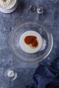 Paprika and garlic powder on top of buttermilk in a glass bowl on a grey counter.