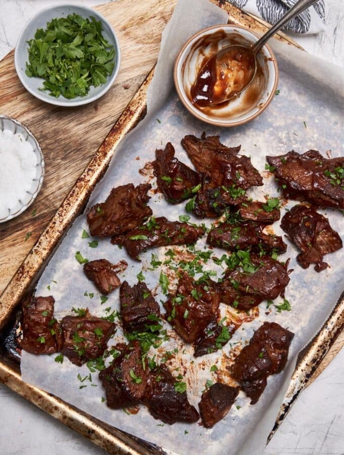 Overhead view of burnt ends garnished with fresh parsley on a baking sheet, next to small bowls of bbq sauce, chopped parsley, and salt.