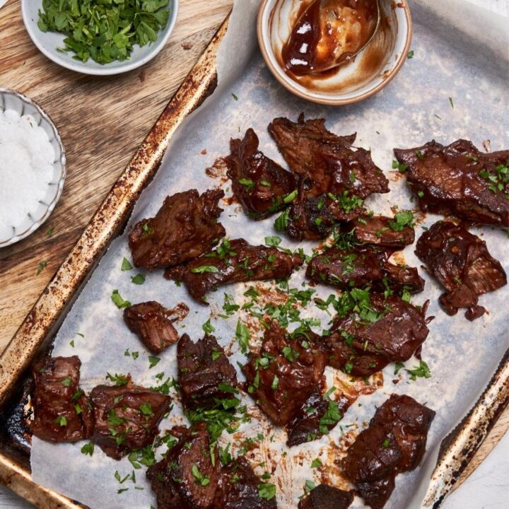 Overhead view of burnt ends garnished with fresh parsley on a baking sheet, next to small bowls of bbq sauce, chopped parsley, and salt.