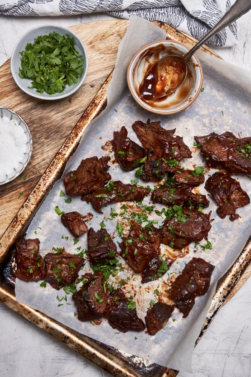 Overhead view of burnt ends garnished with fresh parsley on a baking sheet, next to small bowls of bbq sauce, chopped parsley, and salt.