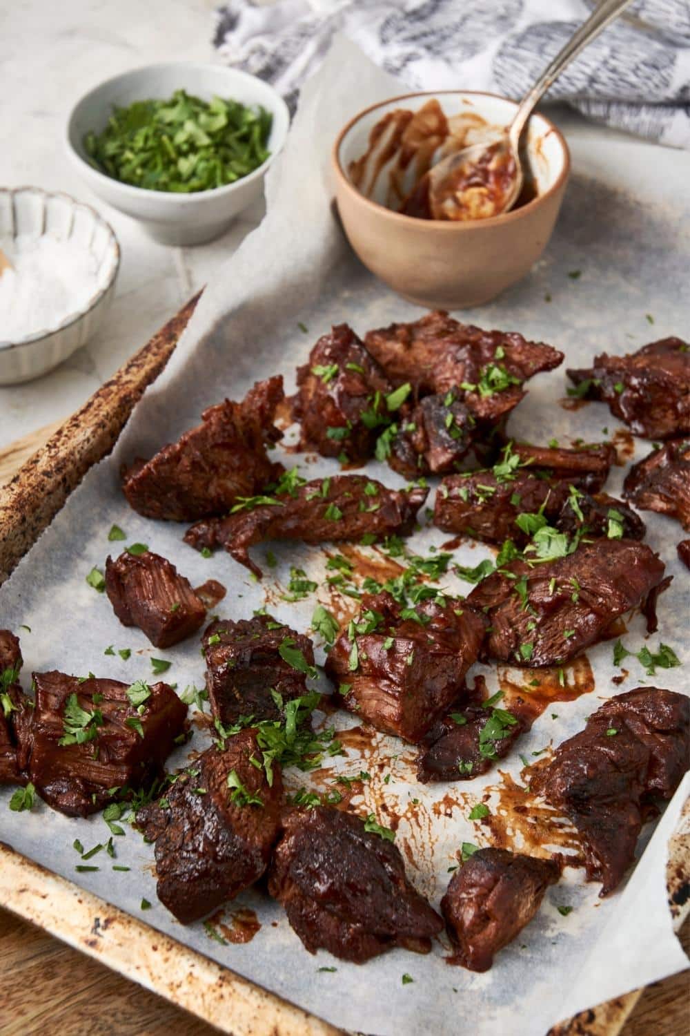 A lined baking sheet with burnt ends topped with fresh parsley, next to small bowls of bbq sauce, herbs, and salt.