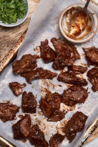 Burnt ends covered with bbq sauce on a baking sheet, next to an emptied bowl of sauce with a spoon.