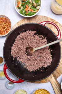 Ground beef in a large red pot with a wooden spoon, surrounded by bowls of vegetables, beef broth, and spices.