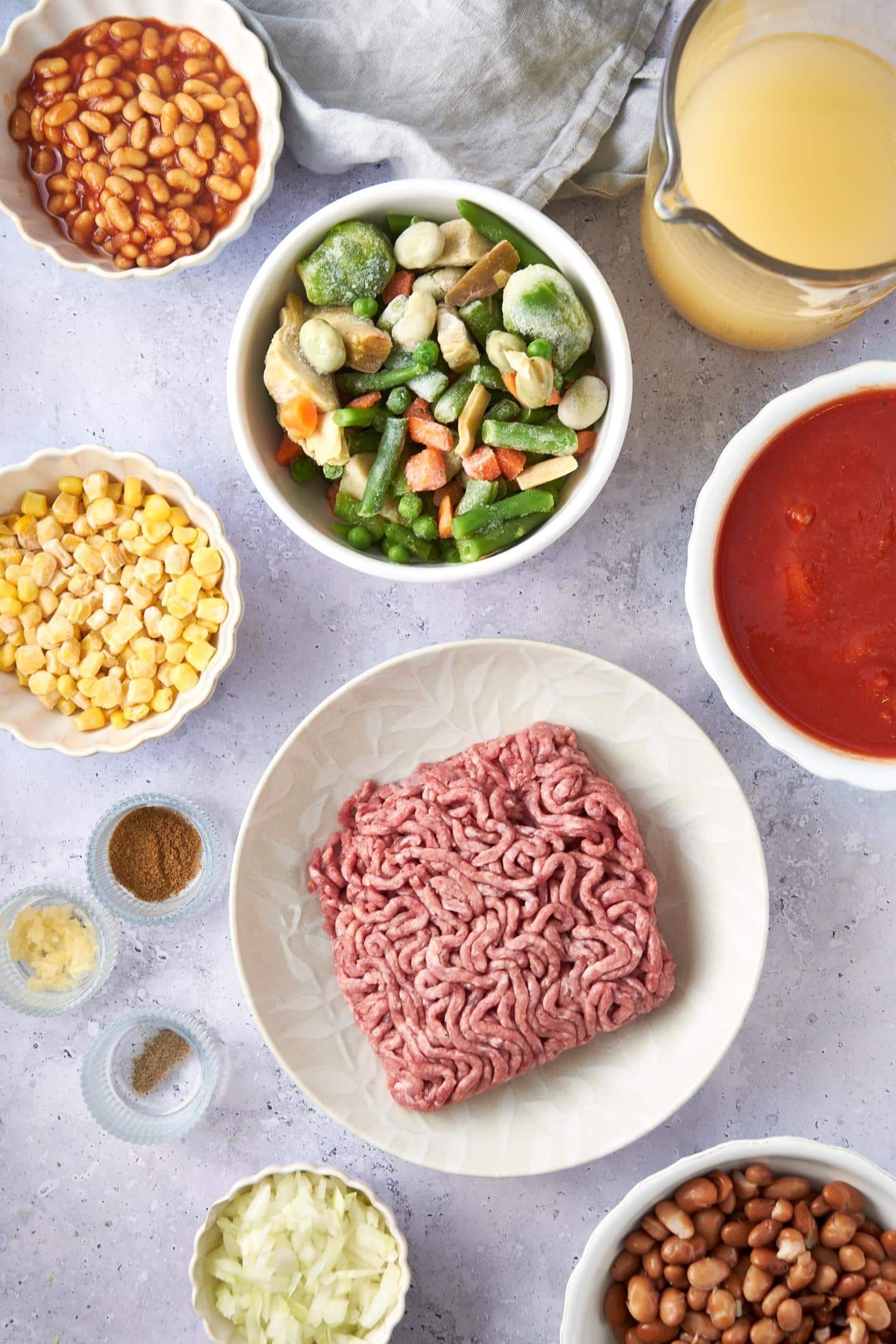 Bowls of ground beef, beef broth, frozen vegetables, beans, corn kernels, diced tomatoes, chopped onions, and spices.