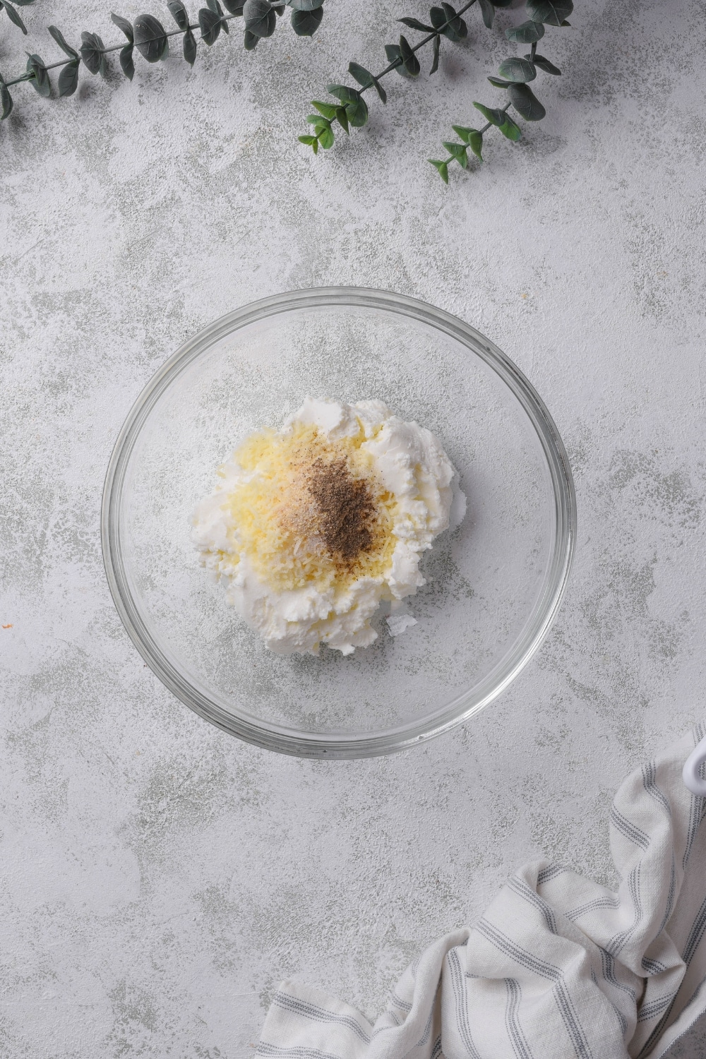 Seasonings and grated parmesan cheese on top of cream cheese in a glass bowl on a grey counter.