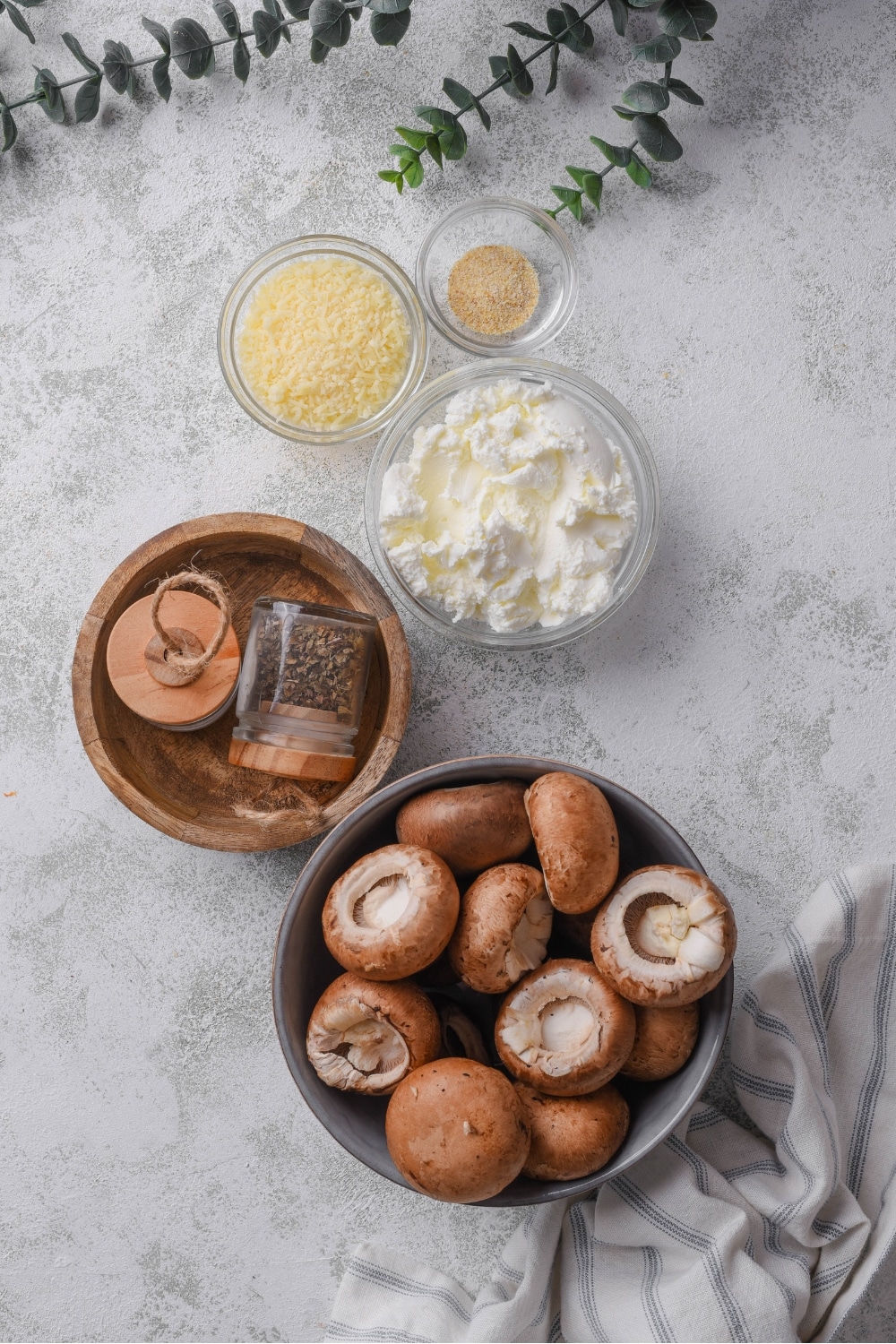 A bowl of mushrooms, a bowl of seasonings in two jars, a bowl of cream cheese, a bowl of parmesan cheese, and a bowl of garlic powder all on a white counter.