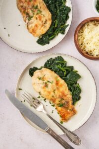 A piece of mushroom chicken on top of collard greens on top of a white plate on a white counter. Behind it is part of a bowl of mozzarella cheese and part of a plate with another piece of chicken on it.