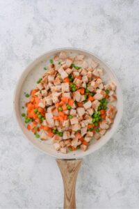 Chopped chicken, carrots, and peas in a white frying pan with a wooden handle.