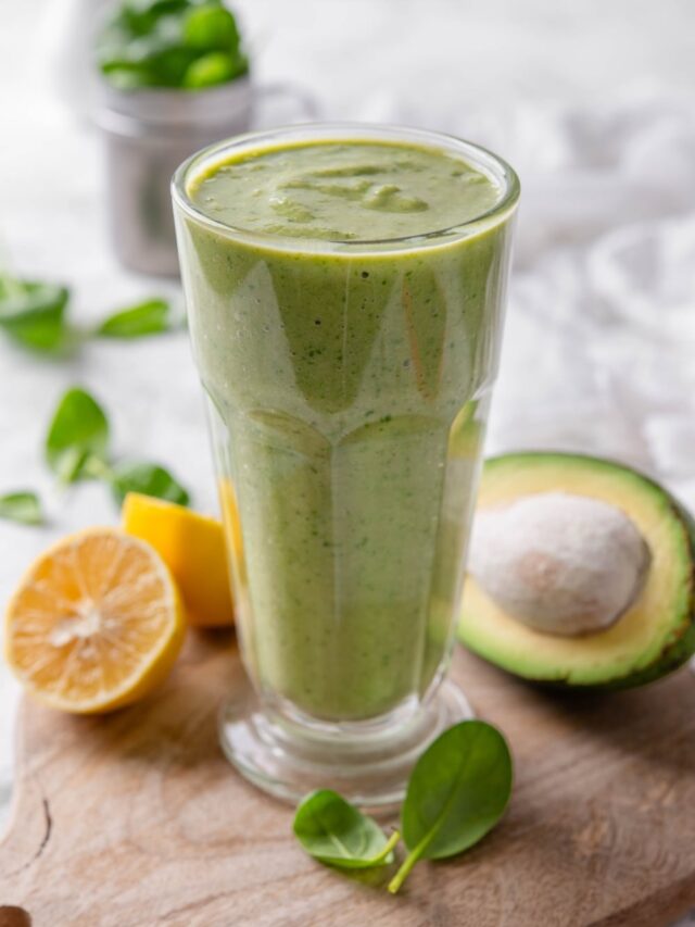 A green smoothie in a tall glass on a wooden board surrounded by spinach leaves, a halved lemon, and a halved avocado.