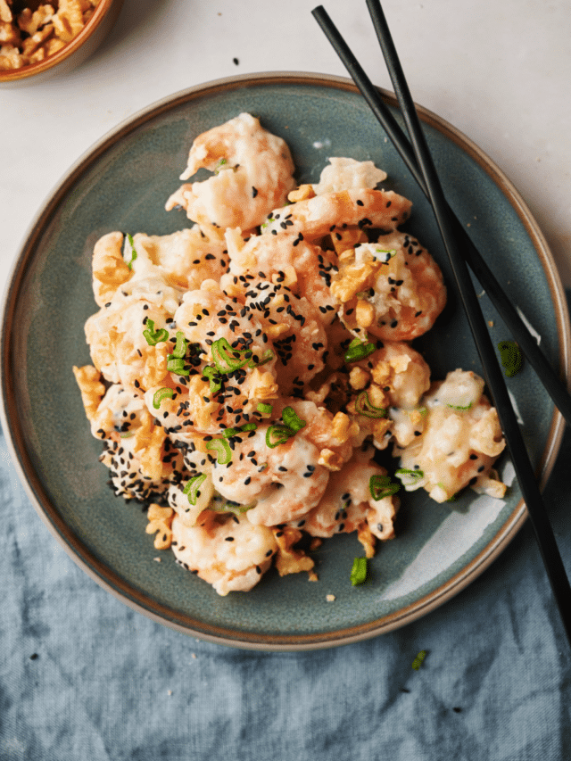 A plate of honey walnut shrimp garnished with sesame seeds, sliced green onions, and walnuts. A set of chopsticks is on the plate.