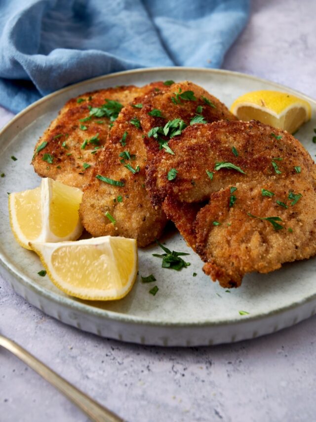 Three breaded turkey cutlets on a plate garnished with fresh herbs and served with lemon wedges.