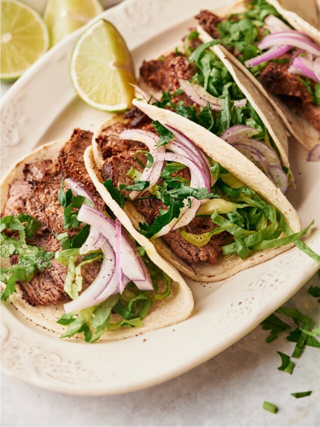 A couple of steak tacos with red onion and cilantro in the tortillas on a white plate.