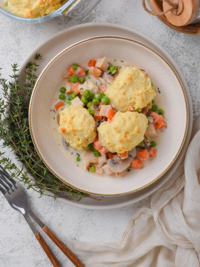 A top-down view of a white bowl full of chicken and biscuits casserole. Three biscuits sit atop the casserole filling. The bowl is sitting on a white plate with a sprig of fresh herbs.