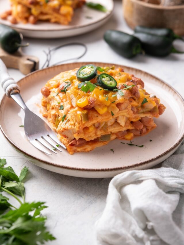 A slice of enchilada casserole with melted cheese, corn, chicken, beans, and peppers in it. The casserole is garnished with two jalapeño slices and fresh herbs and there is a fork on the plate.