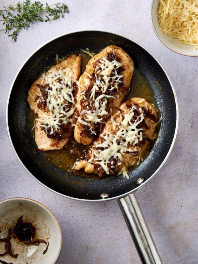A skillet of chicken cutlets in french onion sauce topped with caramelized onions and melted shredded cheese.