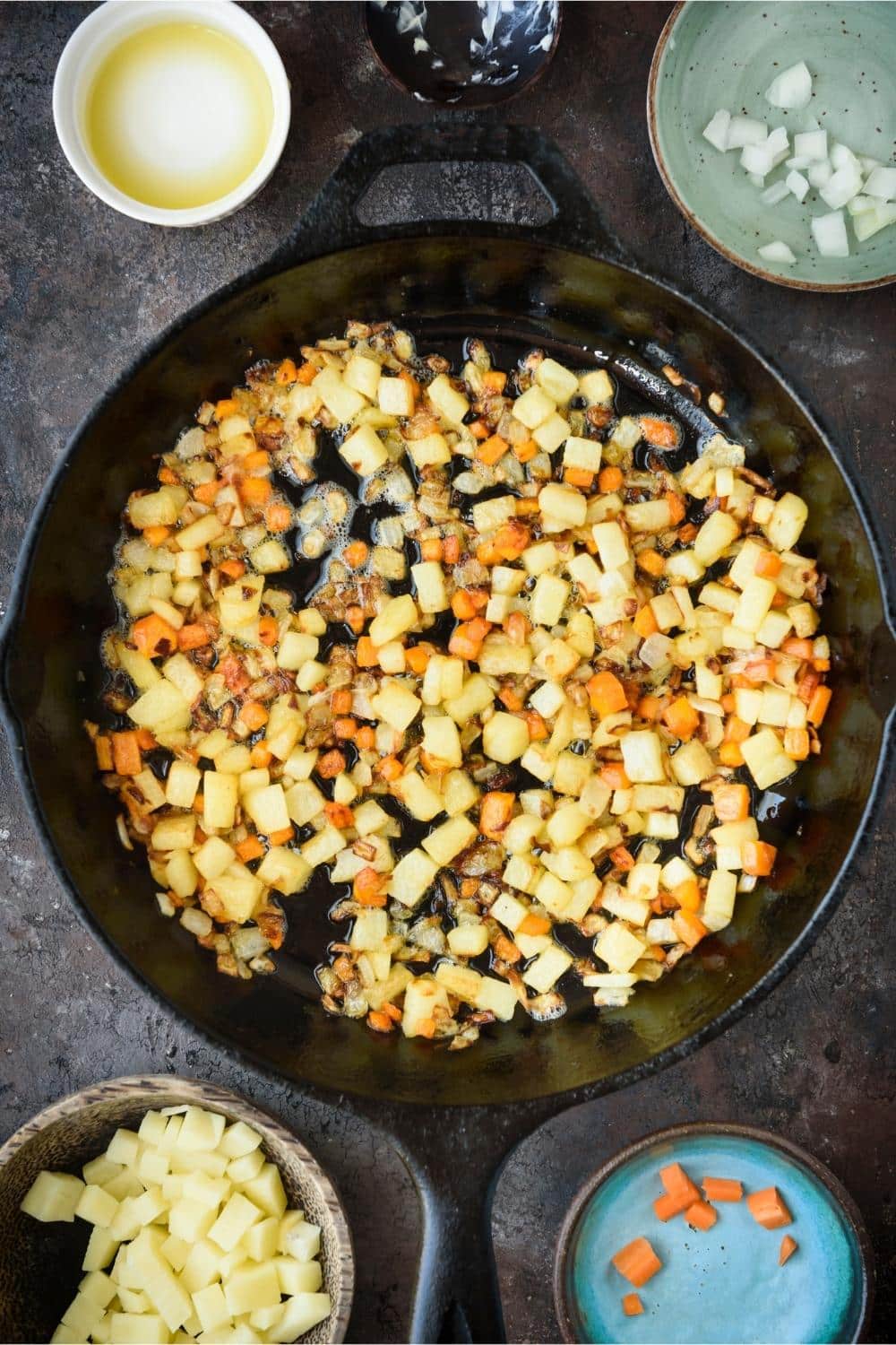 Sauteed potatoes, carrots, and onions in a cast iron pot, surrounded by bowls of extra ingredients.