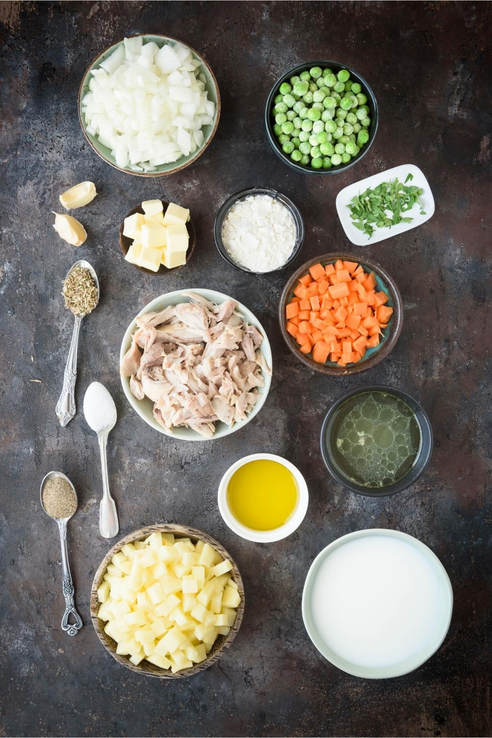 Bowls of shredded cooked chicken, chopped onions, frozen peas, butter, flour, chopped parsley, diced carrots, chicken broth, olive oil, heavy cream, chopped potatoes, teaspoonfuls of spices, and two garlic cloves.